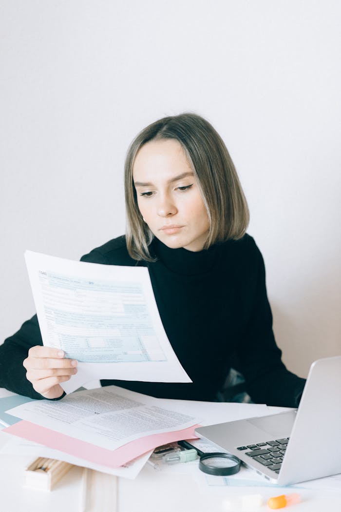 Focused woman reviewing paperwork at her desk with a laptop, showcasing a professional working environment.