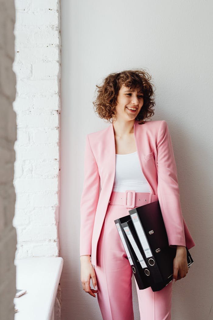 The Art of Drawing Readers In: Your attractive post title goes here Smiling woman in a pink suit holding binders, embodying modern business chic.